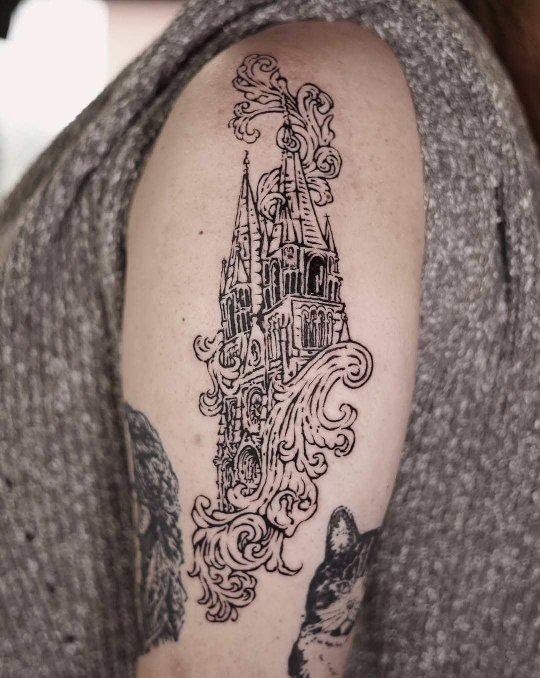 Tatuaż przedstawiający strzelistą katedrę otoczoną dekoracyjnymi ornamentami, wykonany w detalicznym, linorytowym stylu.