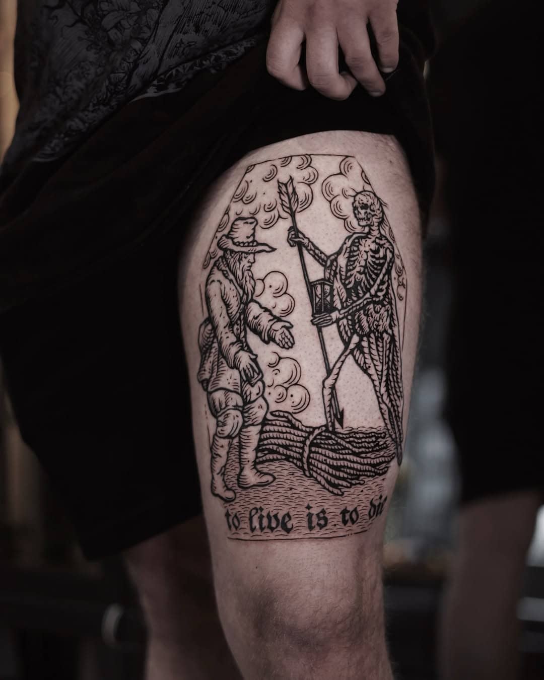 Tatuaż w stylu ryciny przedstawiający scenę „to live is to die” na udzie.