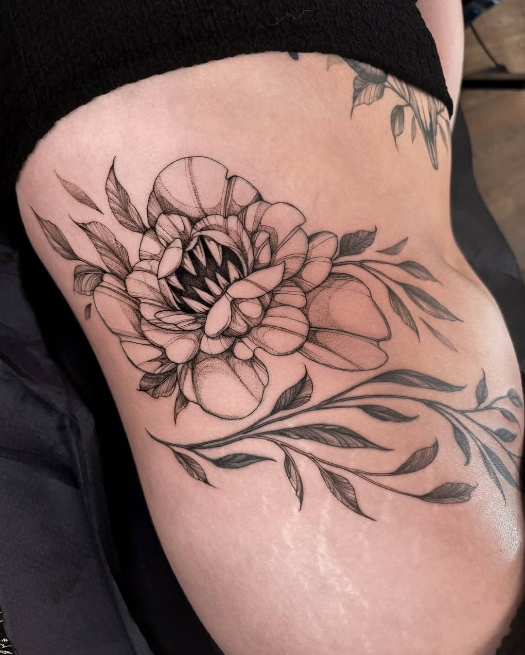 Delikatny tatuaż w stylu fineline przedstawiający kwiat z zębami na udzie – Nava, Sztorm Tattoo Gdańsk