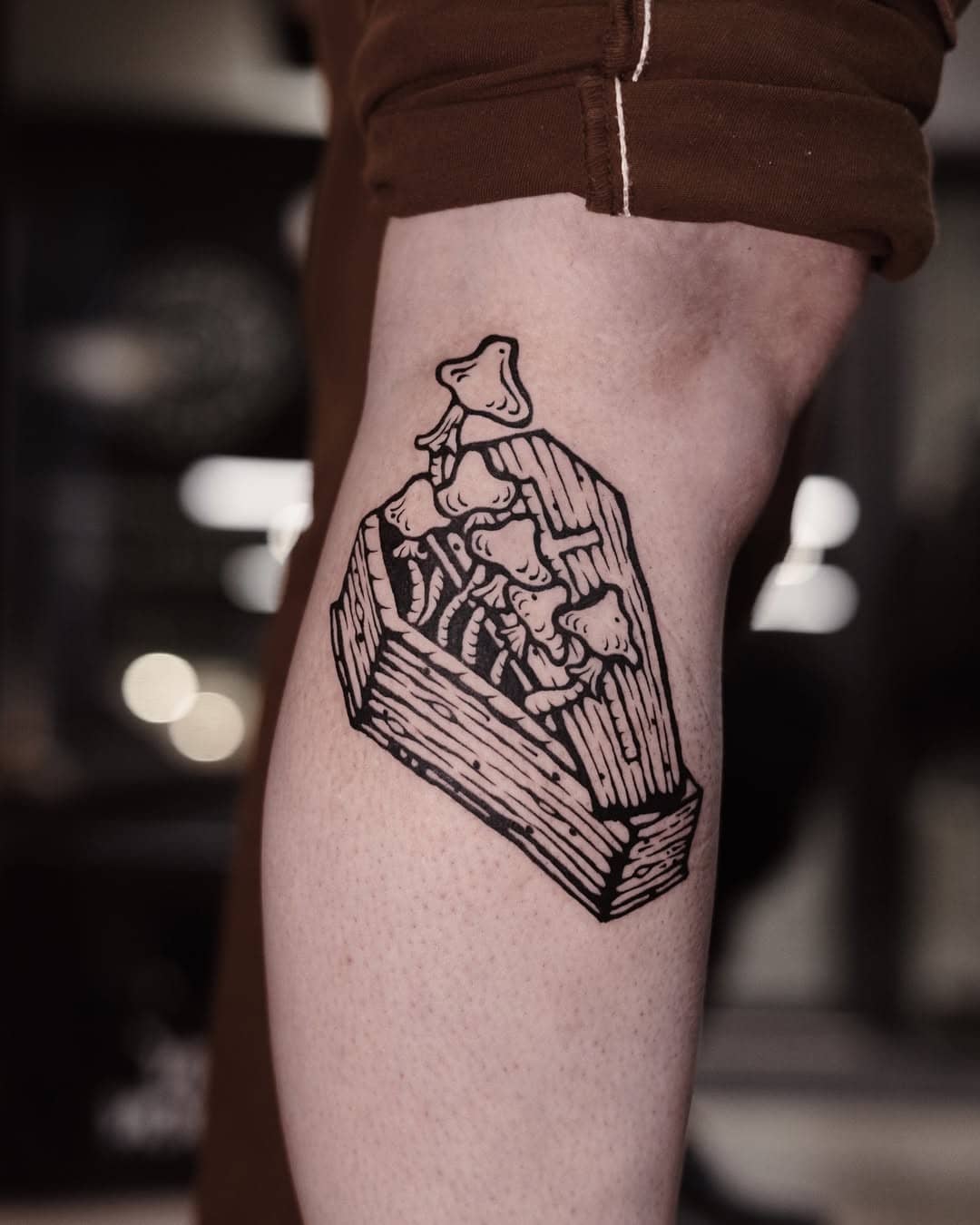 Tatuaż na nodze przedstawiający trumnę, z której wyrastają grzyby, wykonany w linorytowej stylistyce przez Orianę w Sztorm Tattoo Gdańsk.