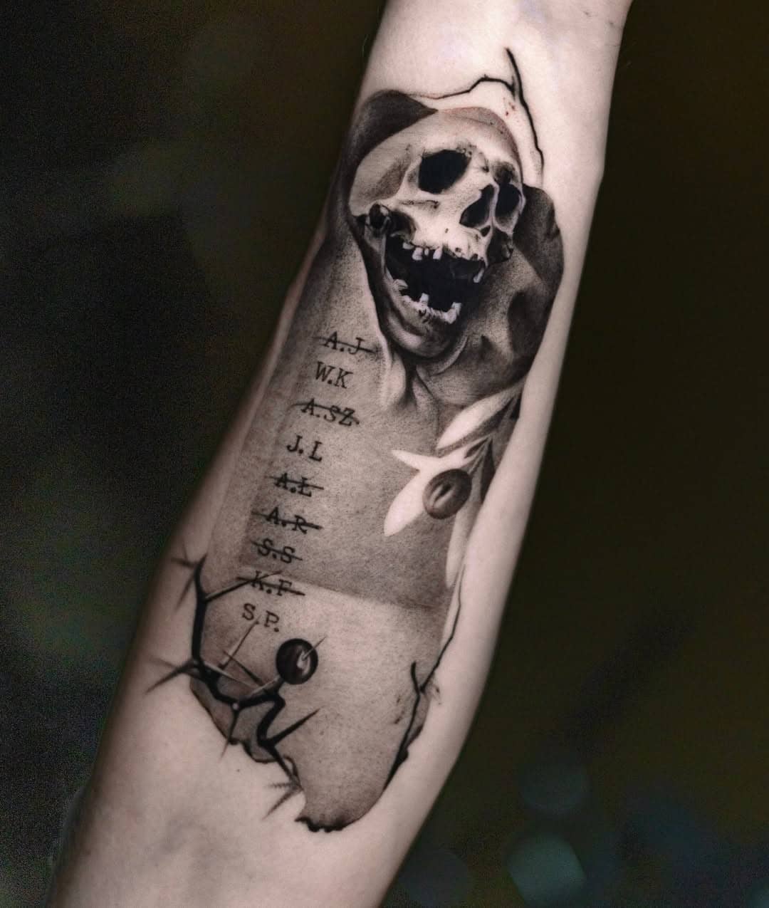 Tatuaż na przedramieniu w stylu realistyczno-graficznym, przedstawiający czaszkę z otwartymi ustami, listę inicjałów oraz elementy dekoracyjne z pęknięciami i kontrastowym cieniem.